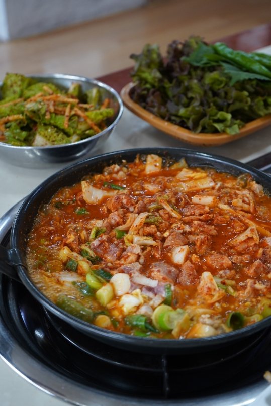 상 위 버너에서 끓는 비빔돼지 냄비, 뒤로 쌈 채소와 겉절이